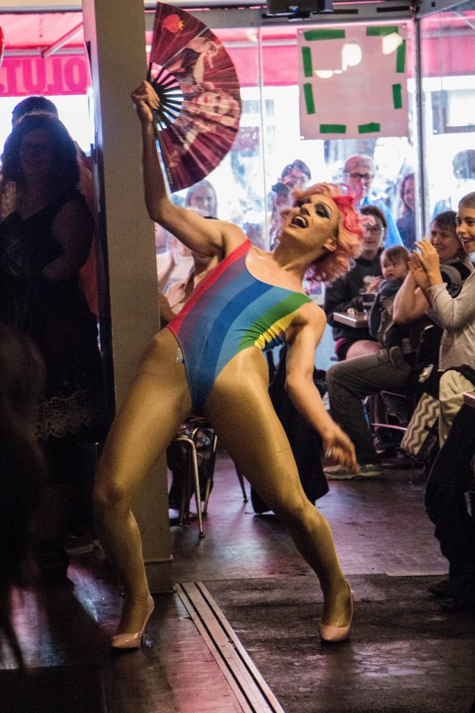 By Asha Collins, Glad Day Bookstore; June 24 - Erin Brockobic performing at Glad Day Bookshop’s weekly Drag Brunch. Erin is wearing a fabulous rainbow leotard, while in a bent-back position with her fan extended. 