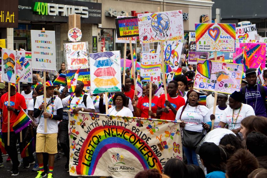 By Asha Collins, Parade Day: June 24 - LGBTQ+ Newcomers group with multiple signs describing how pride accepts all people, and supports newcomers in their journeys starting in Canada. 