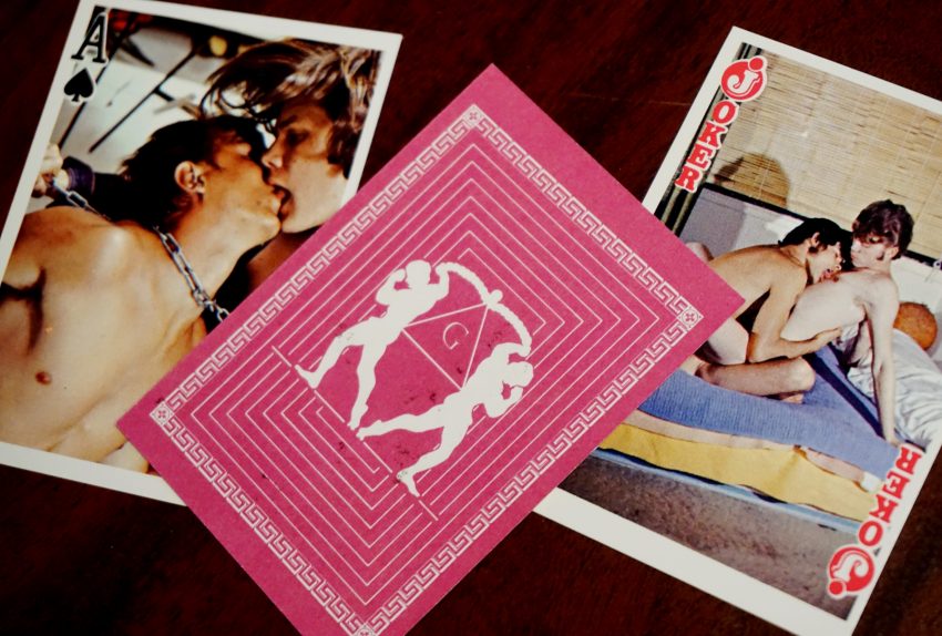 Three playing cards, with one face down in the middle. The face up cards both depict two men kissing while the face down card depicts the silhouettes of two men flexing and facing away from each other atop a graphic pattern.