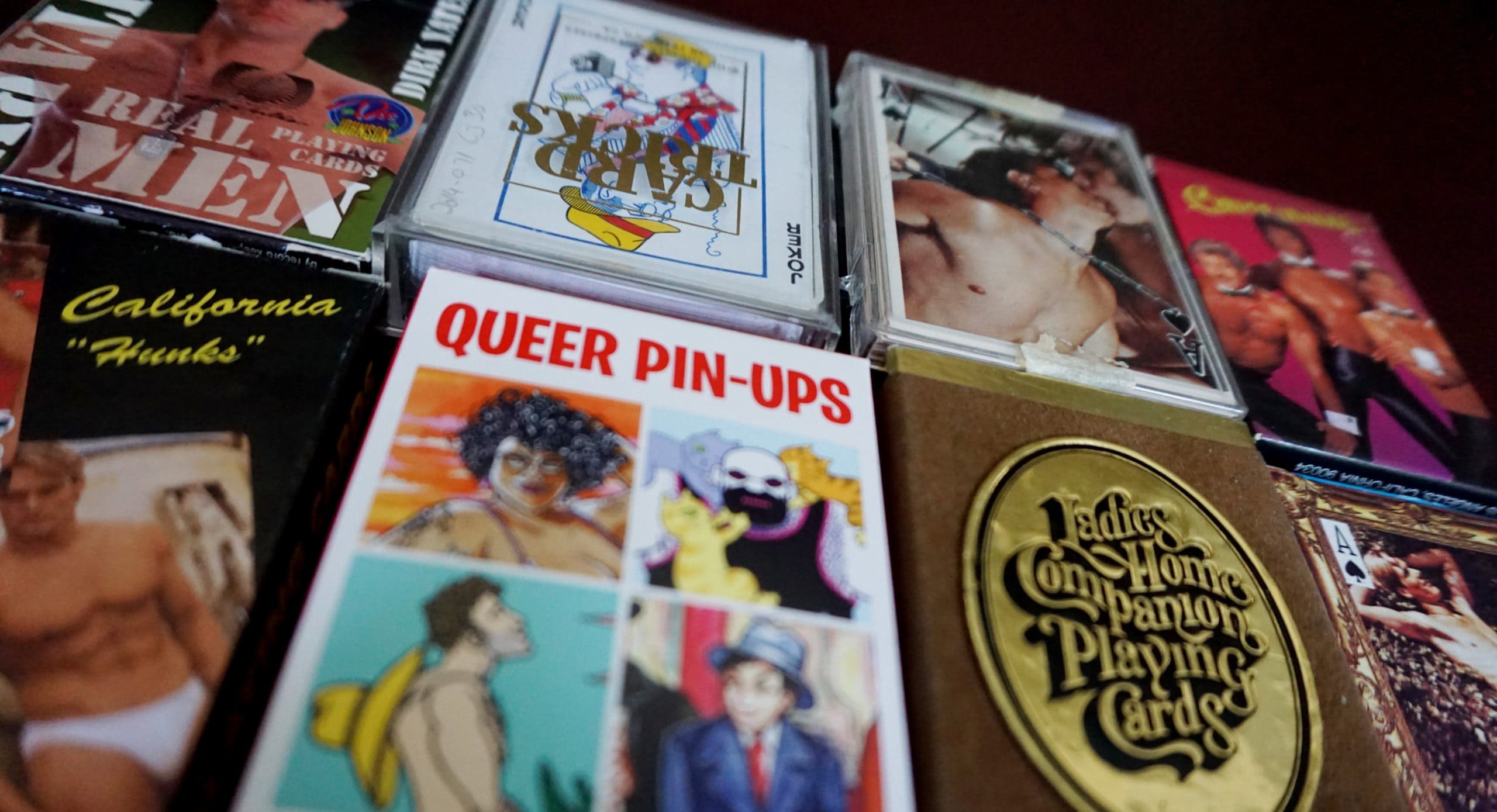 Eight decks of playing cards arranged 4x2 on a table. Queer Pin-Ups and Ladies Home Companion Playing Cards are in focus in the foreground.