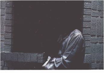 Person in a trench coat against a brick wall with their face in the shadows