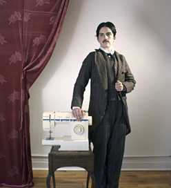 Masculine person in 1900s clothing standing next to a sewing machine.