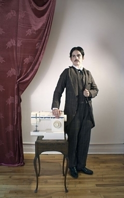 Masculine person in 1900s clothing standing next to a sewing machine.