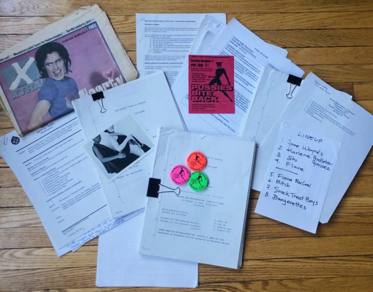 Variety of documents on a wooden table, including an Xtra! newspaper, Pussy Palace button, and a flyer.