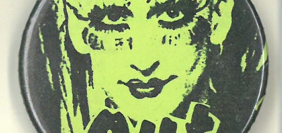 Yellow button with drawing of a person with mascara, eyeliner and lipstick, with the word ‘Out’
