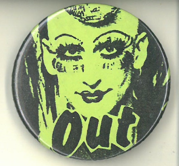 Yellow button with drawing of a person with mascara, eyeliner and lipstick, with the word ‘Out’