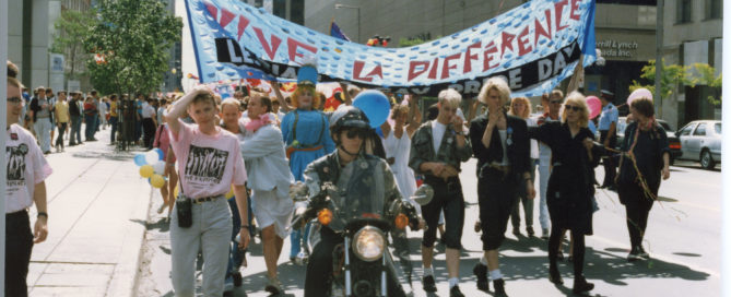 Group parades down street holding “Vive la difference” flag, led by a motorcycle
