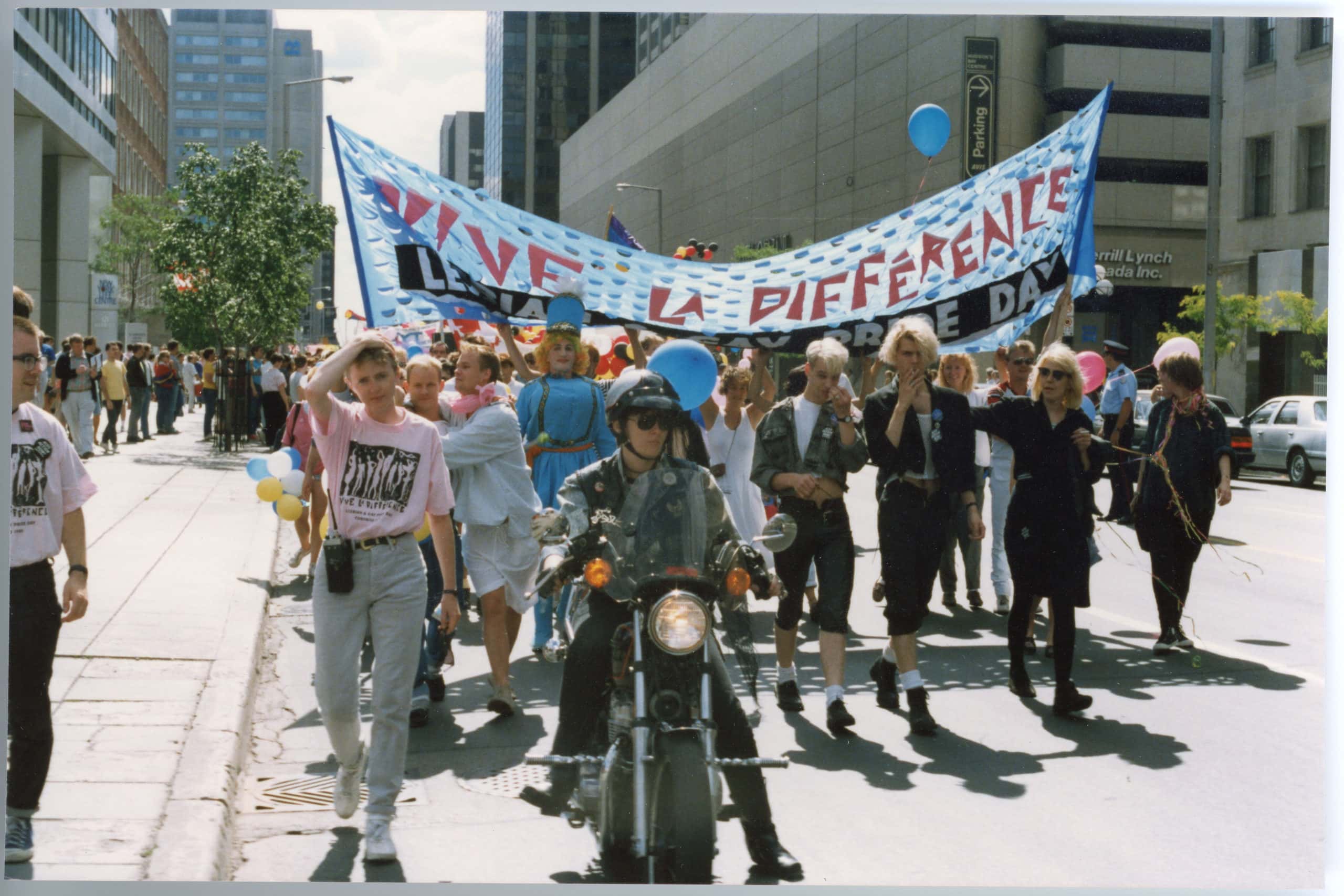 Group parades down street holding “Vive la difference” flag, led by a motorcycle