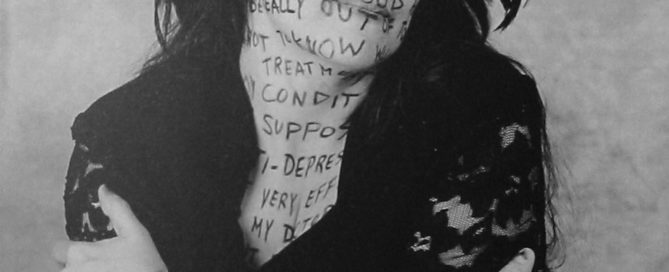 Black and white photo of a person in a hat embracing themselves, with words written on their skin