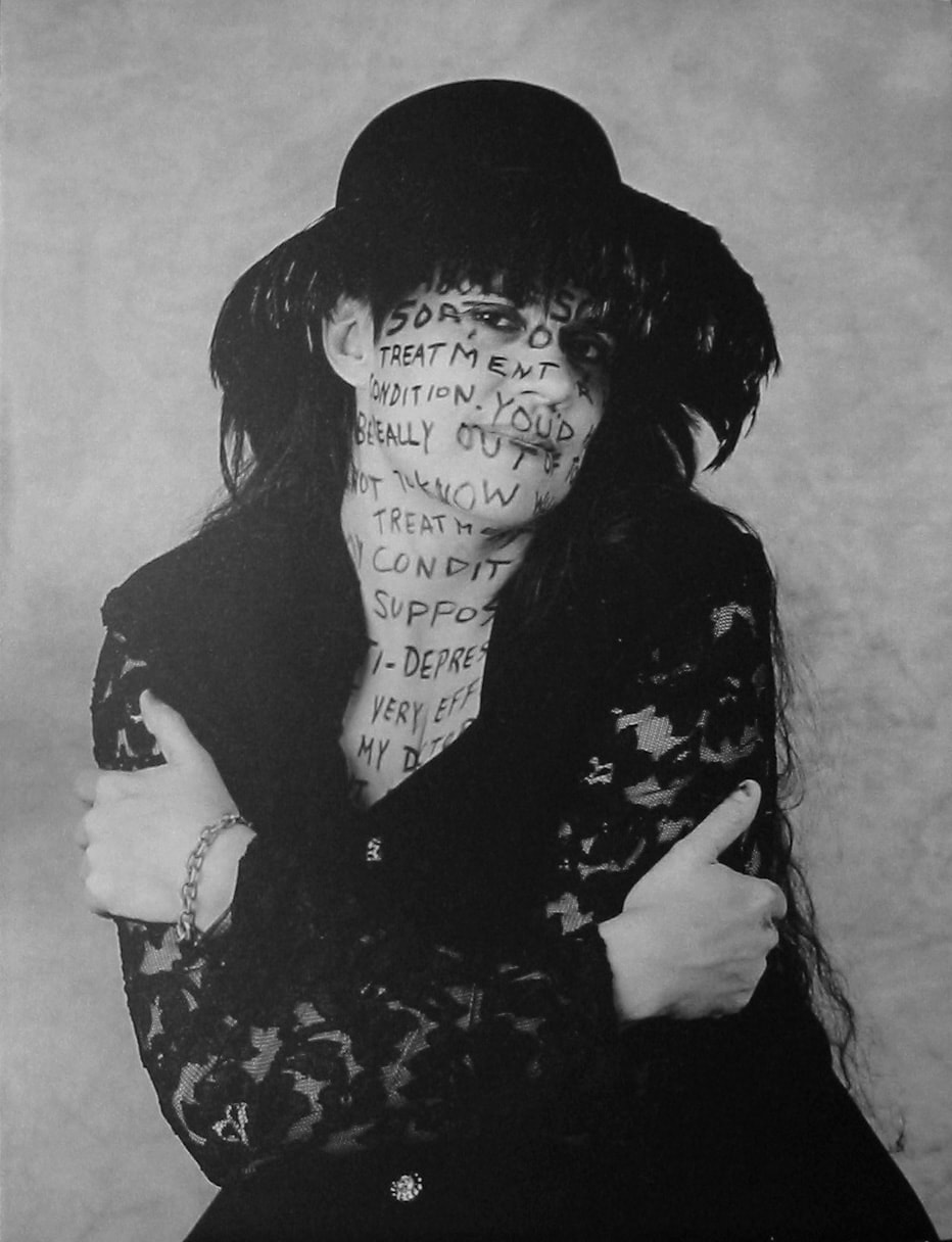 Black and white photo of a person in a hat embracing themselves, with words written on their skin