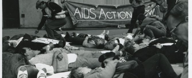 Black and white photo of people lying on the ground, with a photographer taking photos and two people holding a banner reading "AID Action Now"