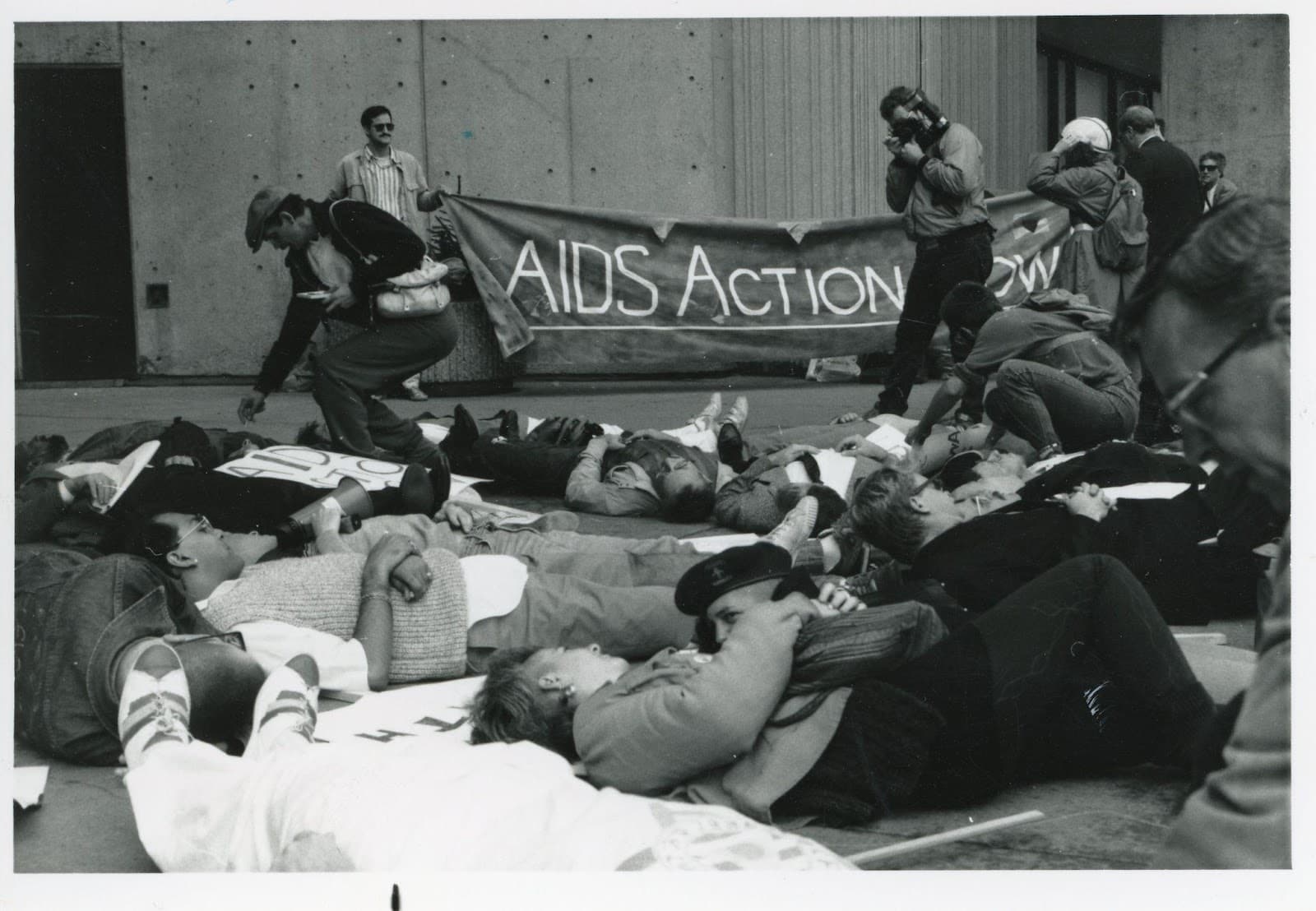 Black and white photo of people lying on the ground, with a photographer taking photos and two people holding a banner reading "AID Action Now"