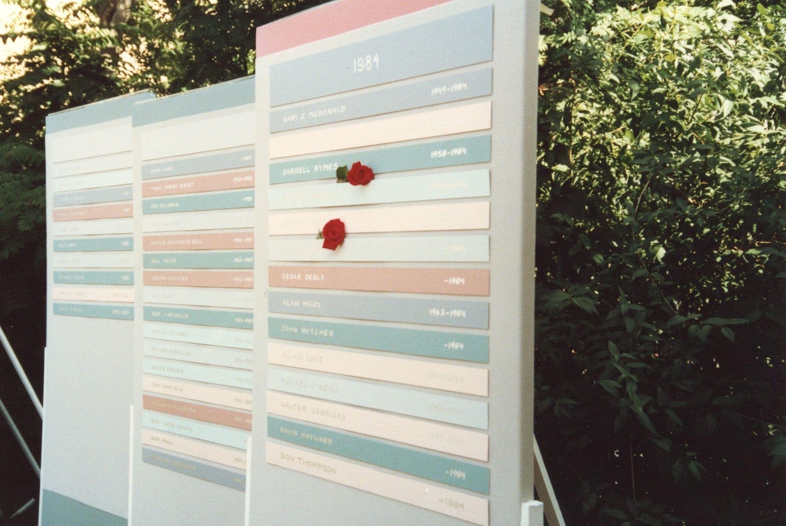 Three panels of an AIDS memorial, labelled ‘1981.’ On each panel, there are a number of rectangular plaques arranged horizontally. Each plaque includes a person’s name and the date of death; some also include dates of birth. The memorial is light grey with accents in pastel pink, blue, green, and cream.