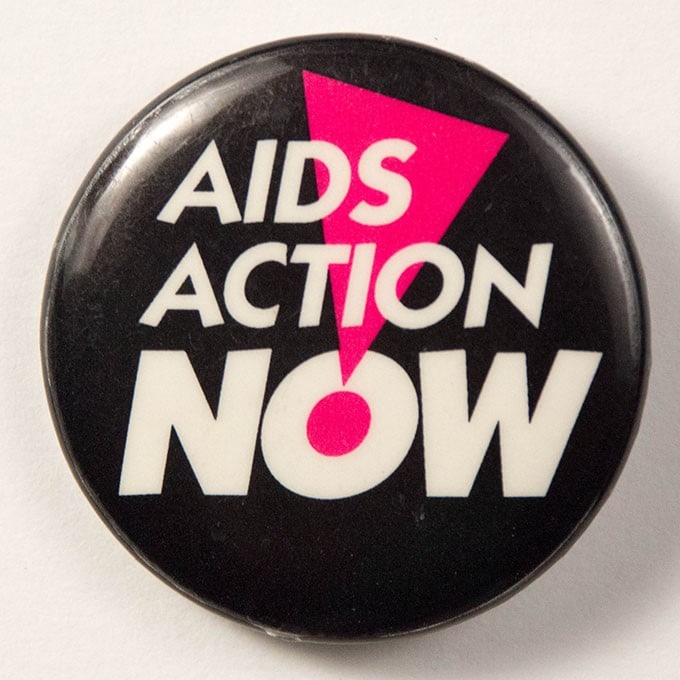 Black button with white text reading, “AIDS Action Now!” in all caps. The exclamation mark is an inverted hot pink triangle with a hot pink dot at the bottom point.