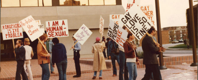 Photo of a group of protestors demonstrating outside the University of Windsor. They are standing in an open area paved with red bricks, and a concrete building with mirrored windows is visible in the background. Some are holding signs reading, “homophobia is the problem,” “reinstate John Damien,” “I am a human-sexual,” “Windsor Gay Unity,” “gay people make it!” and “gay love is so fine!” Others are holding signs that are illegible.