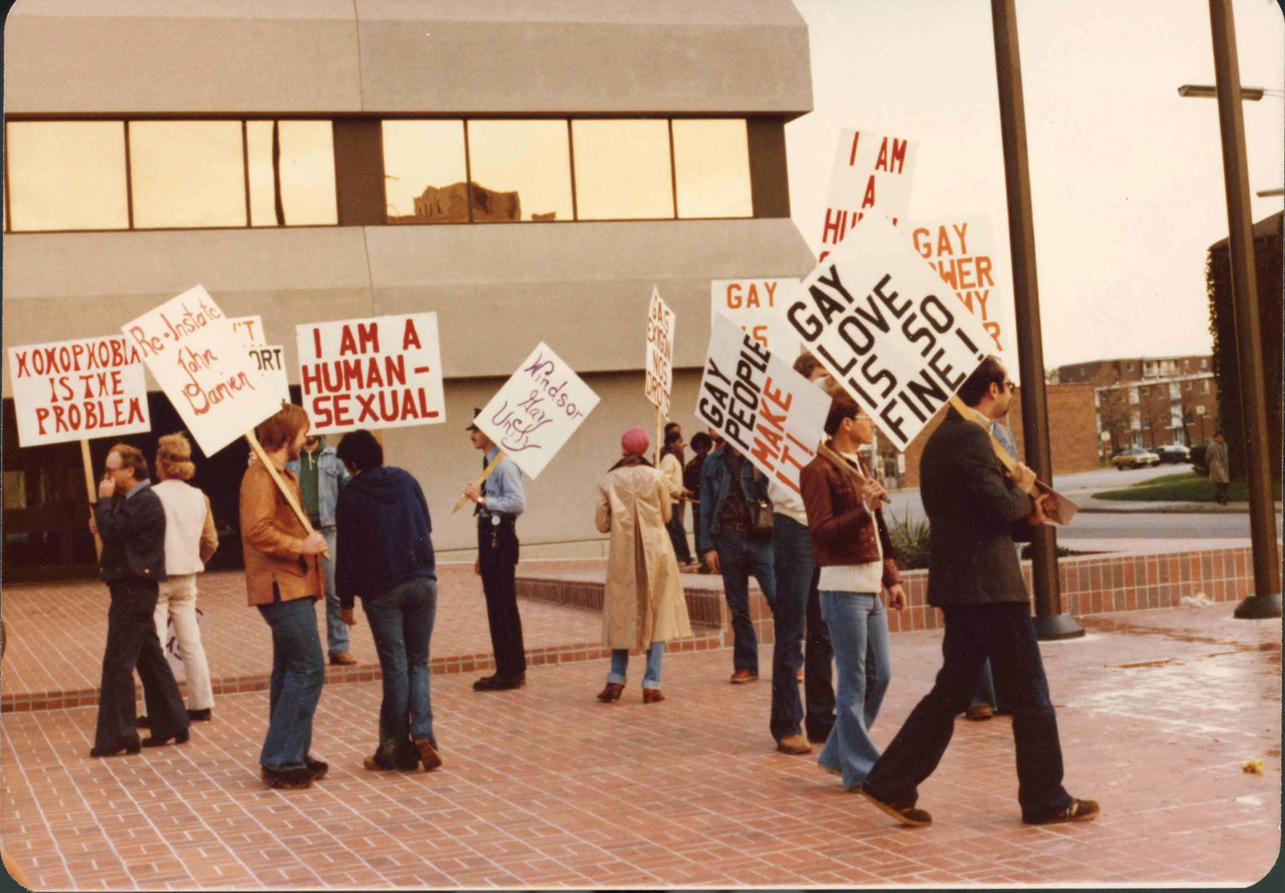 Photo of a group of protestors demonstrating outside the University of Windsor. They are standing in an open area paved with red bricks, and a concrete building with mirrored windows is visible in the background. Some are holding signs reading, “homophobia is the problem,” “reinstate John Damien,” “I am a human-sexual,” “Windsor Gay Unity,” “gay people make it!” and “gay love is so fine!” Others are holding signs that are illegible.