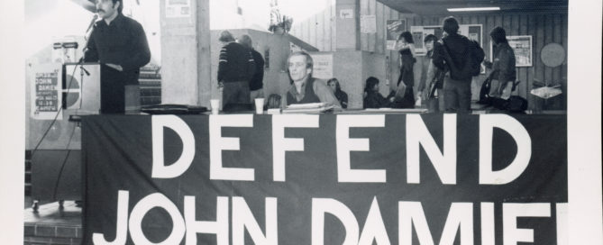 A man with dark sideburns and a moustache is standing at a podium, while another man, dressed in a button-up shirt and sweater vest, sits at a table to his left. A banner reading “defend John Damien” is attached to the front of the table. A number of other people can be seen standing in the background.