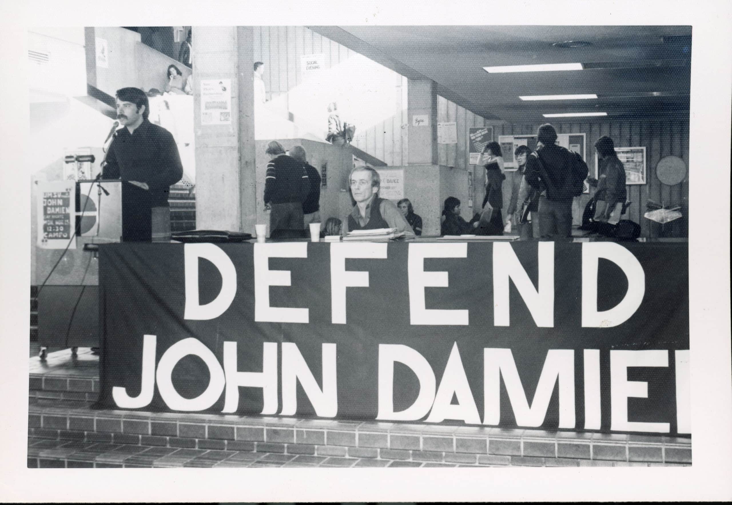 A man with dark sideburns and a moustache is standing at a podium, while another man, dressed in a button-up shirt and sweater vest, sits at a table to his left. A banner reading “defend John Damien” is attached to the front of the table. A number of other people can be seen standing in the background.