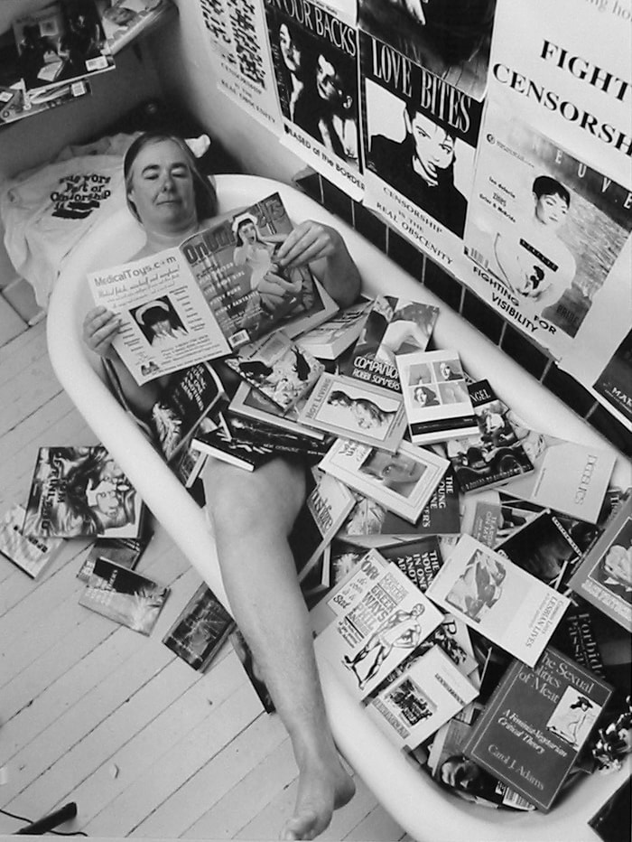 A woman is laying in a large white bathtub with one leg hanging out, reading a magazine. Apart from her right leg, her body is entirely covered by books and magazines; more books and magazines are visible on the floor beside her. There are a number of posters on the wall behind her, including one with the text, “fight censorship.”