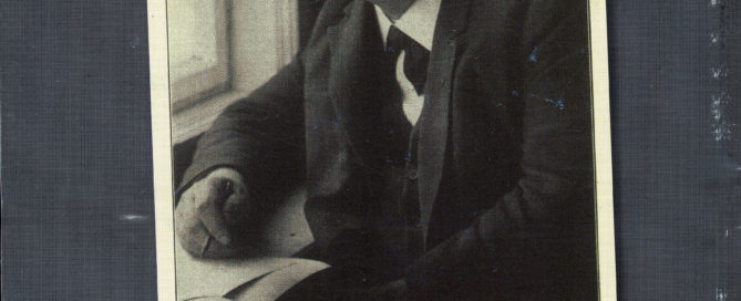 Photo of a middle-aged man seated by a window. He is wearing round, wire-rimmed glasses and a dark coloured suit and is holding a pen in one hand.