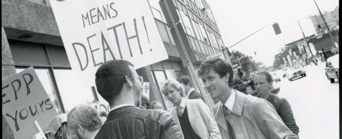 Photo of protestors standing near a building. One is holding a sign reading, “silence means death!” and another is holding a sign reading, “EPP yours.”