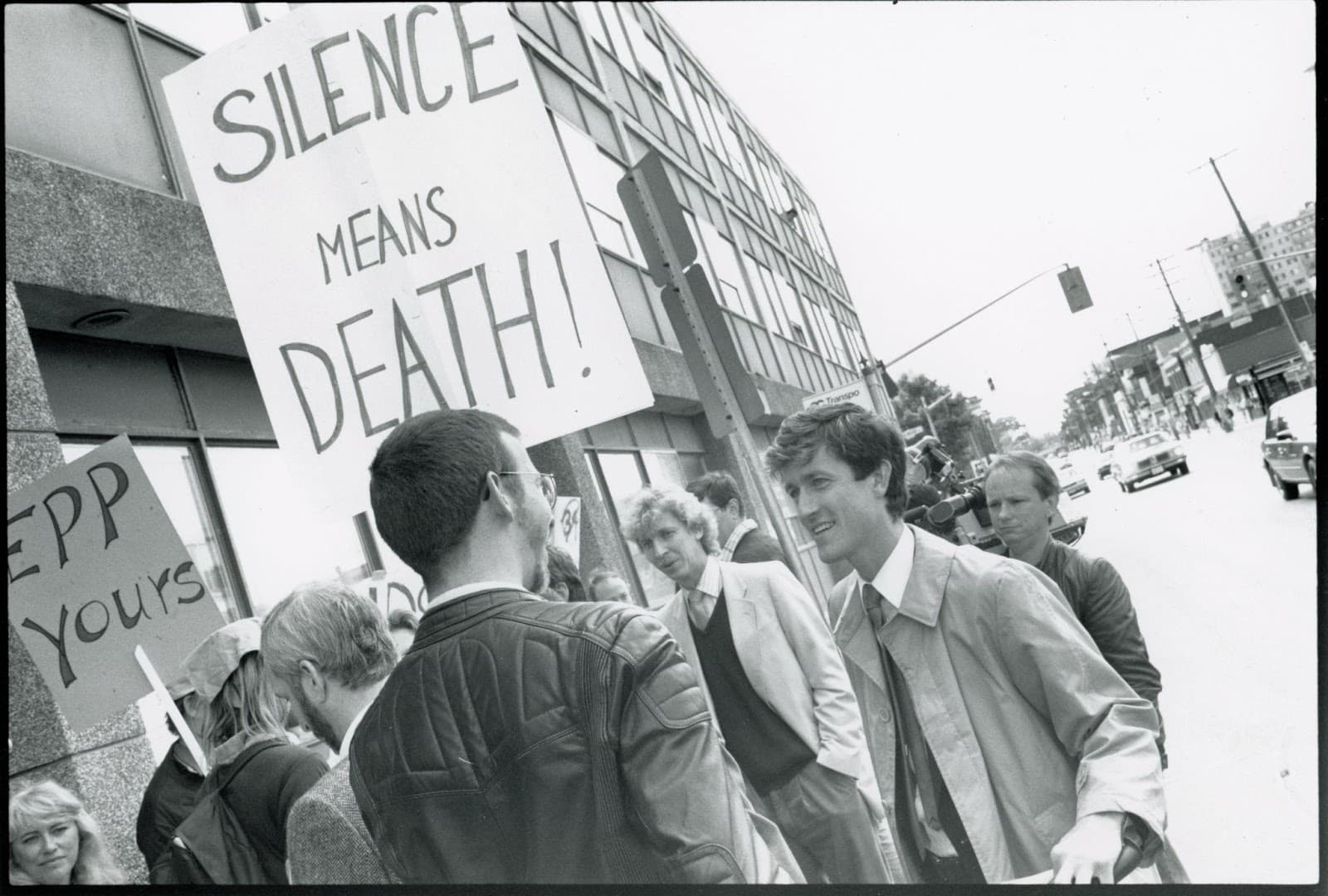Photo of protestors standing near a building. One is holding a sign reading, “silence means death!” and another is holding a sign reading, “EPP yours.”